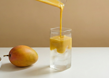 Cremiges Mango-Püree wird in ein hohes eckiges Glas mit Eiswürfeln und Kokoswasser gegossen, daneben eine frische Mango und eine Schale mit Mangopüree auf hellem Hintergrund – tropisches Sommergetränk mit Mango und Kokos.