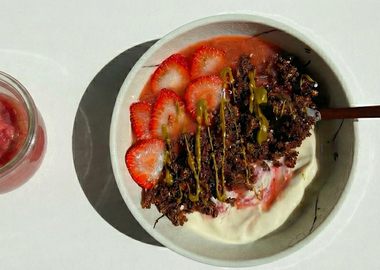 Veganes Erdbeer-Rhabarber-Kompott im Glas neben einer Bowl mit pflanzlichem Joghurt, frischen Erdbeeren, Schoko-Granola und Nussmus als Topping.