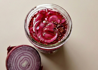 Glas mit pink eingelegten roten Zwiebelringen in Essig Sud mit Senfsamen und Koriandersamen, daneben eine halbierte rote Zwiebel auf hellem Hintergrund – selbstgemachte Pickled Onions.