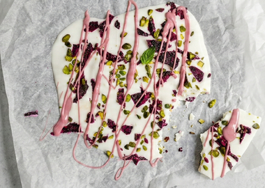 Gefrorener Joghurt auf einem mit Backpapier belegten Blech, getoppt mit getrockneten Drachenfruchtscheiben, Pistazien, frischer Minze und geschmolzener Ruby-Schokolade.