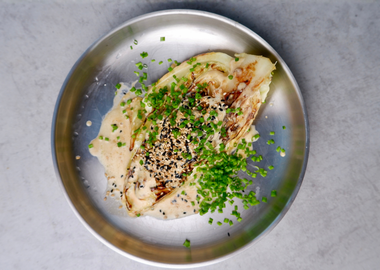 Gebratener Spitzkohl mit cremiger Miso-Rahm-Soße, garniert mit schwarzem und hellem Sesam sowie frischem Schnittlauch auf einem Metallteller angerichtet.