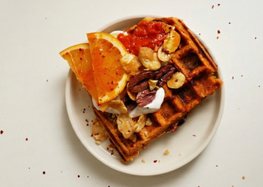 Herzhafte Orange-Chili-Waffel mit Orangenscheiben, veganem Joghurt, Oliven, Chili-Flocken und gerösteten Favabohnen auf einem weißen Teller.