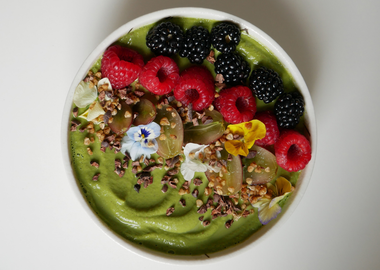 Vegane und glutenfreie grüne Smoothie Bowl aus Avocado, Spinat, Kiwi und Brokkoli, dekoriert mit Himbeeren, Brombeeren, Trauben, Kakaonibs und essbaren Blüten.