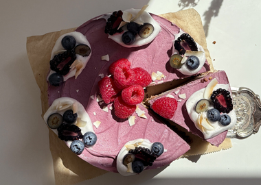 Draufsicht einer runden, lilafarbenen Cashew-Eistorte auf Backpapier. Ein schmuckvoller Tortenheber aus Silber hebt ein angeschnittenes Stück an. Die Torte ist mit Tupfen aus weißer Aquafaba-Sahne, frischen Heidelbeeren, Brombeeren, Himbeeren und Kokoschips garniert. Im Zentrum liegt ein Häufchen roter Himbeeren auf der cremig-gefrorenen Oberfläche.