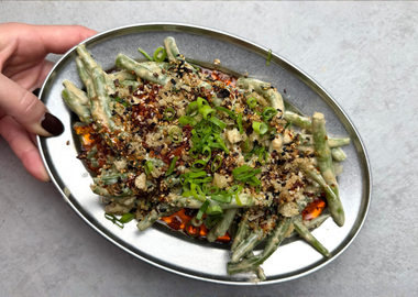 Grüne Bohnen mit cremiger Tahini-Sauce, Chili-Öl, Panko-Brösel, schwarzem Sesam und Frühlingszwiebeln auf einer Servierplatte.