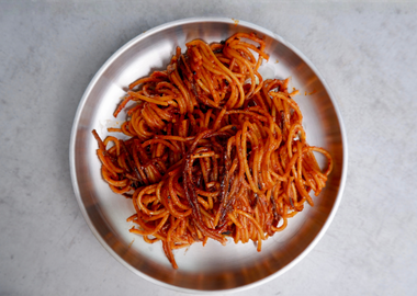 Portion Spaghetti al'assasina in einer glänzenden, tiefroten Tomatensauce, leicht angeröstet und feinen Chiliflocken, serviert auf einem silbernen Metallteller vor einem hellgrauen Hintergrund.