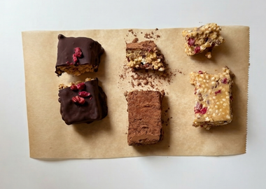 Draufsicht auf drei verschiedene Variationen von hausgemachten Crispy-Quinoa-Vanille-Protein-Riegeln auf braunem Backpapier. Links zwei mit dunkler Zartbitterschokolade überzogene Riegel, garniert mit getrockneten Granatapfelkernen. In der Mitte zwei in Kakaopulver gewälzte Riegel. Rechts zwei Riegel im Naturzustand, die die helle Textur aus gepufftem Quinoa, Soja-Crispies und roten Granatapfelkernen zeigen.