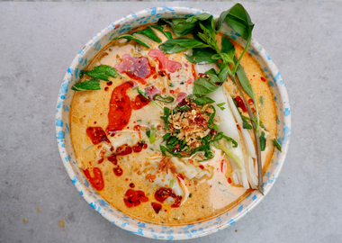 Schale mit cremiger Kokos-Curry-Dumpling-Suppe, gefüllt mit Gyoza und Pak Choi in aromatischer Brühe aus Kokosmilch, roter Currypaste, Ingwer und Knoblauch, garniert mit Frühlingszwiebeln, Röstzwiebeln, Chiliöl und frischen Kräutern auf hellem Hintergrund.