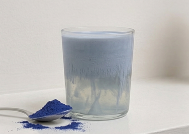 Ein Glas mit einem hellblauen, schaumigen Kokosdrink steht auf einer weißen Oberfläche vor einer schlichten Wand, mit leichter Kondensation am Glas. Daneben liegt ein Löffel mit intensiv blauem Spirulina-Pulver, von dem etwas auf die Oberfläche gestreut ist. Im Hintergrund ist unscharf eine Verpackung mit der Aufschrift „Kokos Wasser“ zu sehen.