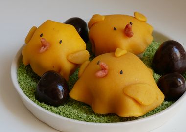 Drei kleine, runde Osterküken-Kuchen aus gelbem Marzipan mit rosa Schnäbeln und schwarzen Augen aus Sesam. Die Küken sitzen auf grün gefärbten Kokosraspeln in einer weißen Schale, dekoriert mit dunklen Schokoladeneiern auf weißem Hintergrund.