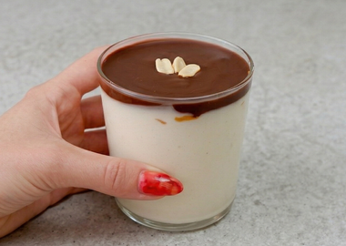 Ein cremiges Protein-Erdnuss-Dessert in einem Glas, serviert auf einer minimalistischen, grauen Arbeitsplatte. Das Dessert besteht aus einer hellen Vanille-Protein-Schicht, getoppt mit einer glatten, dunklen Schokoladenglasur und garniert mit drei Erdnüssen in der Mitte. Eine Hand mit rot lackierten Fingernägeln hält das Glas seitlich fest.