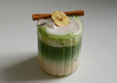 Ein Glas auf einem weißen Tisch, gefüllt mit einem Getränk. Unten ist eine milchige Flüssigkeit, darüber eine Schicht aus leuchtend grünem Matcha-Tee. Das Getränk ist mit Eiswürfeln versehen und hat eine Schaumkrone, die bis über den Rand läuft. Auf dem Schaum liegen eine Zimtstange und eine getrocknete Bananenscheibe.