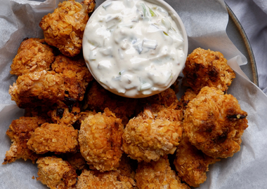Eine Schüssel mit goldbraun gebackenen, knusprigen Blumenkohl-Nuggets, die auf weißem Papier in einer Schale angerichtet sind. Darin steht eine kleine weiße Schale mit einem cremigen Kräuter-Dip. 