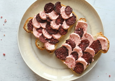 Zwei Scheiben geröstetes Brot auf einem hellen Teller, belegt mit rosa aufgeschlagener veganer Butter und dunklen Rote-Bete-Chips in einem dekorativen Schuppenmuster auf einer Marmoroberfläche.