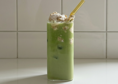 Ein hohes Glas mit grünem Matcha-Bananen-Pudding Drink und gelbem Strohhalm vor einer weißen Fliesenwand.