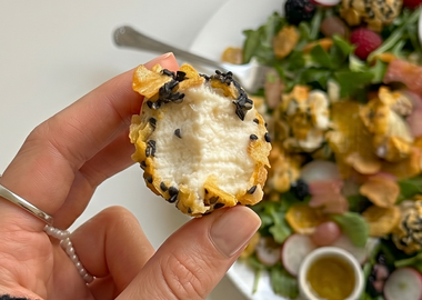 Nahaufnahme einer Hand, die ein knusprig paniertes Air Fryer Tofu 'Veta' Bite mit schwarzem Sesam hält, vor einem Hintergrund mit frischem Salat auf einer weißen Ablage.