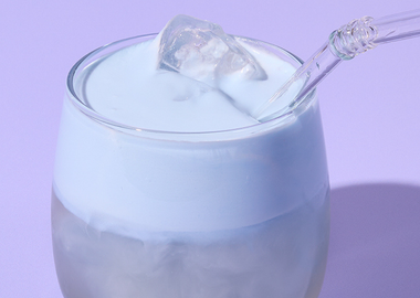Cremiger Blue Spirulina Coconut Cloud Drink mit Eiswürfeln und Glasstrohhalm vor lila Hintergrund – modernes Trendgetränk.