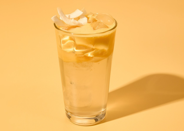 Erfrischender Mango Coconut Cloud Water Drink mit Eiswürfeln und Kokoschips in einem Glas vor gelbem Hintergrund – tropisches Sommergetränk.