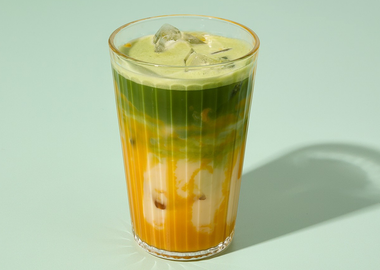 Grüner Matcha-Mango Latte mit Milch und Eiswürfeln im Glas vor mintfarbenem Hintergrund – iced Matcha-Getränk in hochwertiger Produktfotografie.