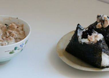 Zwei vegane Tuna-Mayo-Onigiri umwickelt mit Nori-Alge auf einem Teller neben einer Schüssel mit veganer Thunfisch-Füllung.