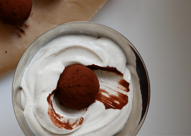 Tiramisu Energy Balls: Ein mit Kakaopulver bestäubtes Energiebällchen liegt eingebettet in cremiger weißer Creme in einer silbernen Schale, weitere Bällchen im Hintergrund auf Backpapier.