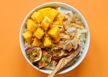 Cremige Frühstücks-Bowl mit Milchreis, frischen Mangowürfeln, Passionsfrucht und gerösteten Kokoschips mit einem Holzlöffel auf hellorangem Hintergrund.