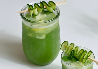 Zwei Gläser erfrischender grüner Gurke-Kokos-Aloe-Refresher, garniert mit aufgespießten Gurkenbändern, auf weißem Hintergrund.