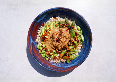 ine Aufnahme einer Schüssel mi braunem Reis, belegt mit geraspeltem Tofu, grünem Spargel, Avocado, Edamame, einer Erdnusssauce und Sesam. Serviert in einer blauen Schüssel auf grauem Hintergrund.