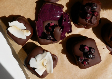 Draufsicht auf fünf Mango-Matcha-Ube-Eis-Bites, überzogen mit Schokolade und garniert mit Kokosraspeln und Johannisbeeren, arrangiert auf Backpapier im Sonnenlicht.