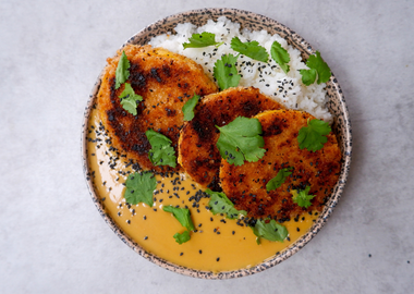 Zentrierte Draufsicht auf eine gesprenkelte Schüssel mit knusprig panierten Butternut-Kürbis-Scheiben, serviert mit weißem Reis und einer cremigen, gelborangen Currysauce. Das Gericht ist mit frischen Korianderblättern und schwarzem Sesam garniert und steht auf einem schlichten, hellgrauen, leicht strukturierten Untergrund im Querformat.