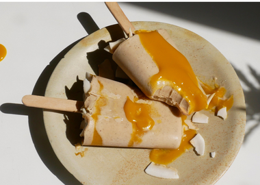 Zwei vegane Mango-Vanille Popsicles auf einem rustikalen Teller in hellem Sonnenlicht, großzügig übergossen mit fruchtigem Mangopüree und garniert mit gerösteten Kokoschips.