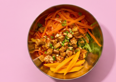 Eine Edelstahlschüssel mit Glasnudelsalat, garniert mit gebratenen Chili-Tofu-Würfeln, frischen Mangostreifen, Karotten und Erdnüssen, fotografiert von oben auf einem rosa Hintergrund.