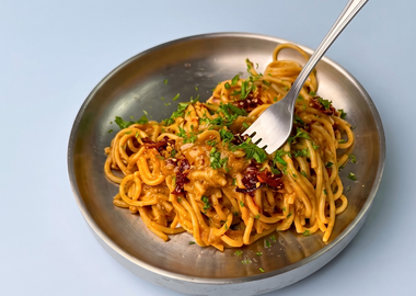 Ein Metallteller mit veganer Pasta mit karamellisierten Zwiebeln, garniert mit frischen Kräutern und getrockneten Tomatenstücken, in die eine Kuchengabel hineinsticht, fotografiert vor einem hellblauen Hintergrund.Ein Metallteller mit veganer Pasta mit karamellisierten Zwiebeln, garniert mit frischen Kräutern und getrockneten Tomatenstücken, in die eine Kuchengabel hineinsticht, fotografiert vor einem hellblauen Hintergrund.
