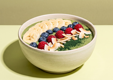 Eine hellgrüne Spriulina-Bowl in einer beigen Keramikschüssel, ordentlich garniert mit Bananenscheiben, Blaubeeren, Himbeeren und Kokoschips vor einem minimalistischen, grünen Hintergrund.