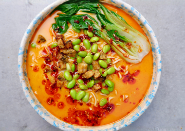 charfe Kokos-Ramen mit gebratenem Tempeh, grünem Gemüse und Chili-Öl in einer gesprenkelten Keramikschüssel.