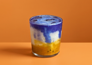 Ein geschichteter Mango-Passionsfrucht-Blue-Spirulina-Latte in einem klaren Glas auf einem warmen, orangefarbenen Hintergrund.