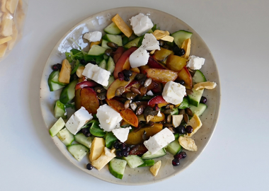 Draufsicht auf einen Salat mit gegrillten Pfirsichen, veganem Feta, Gurken und Kürbiskernen, bestreut mit gefriergetrockneten Mangostücken; oben links steht eine transparente Tüte mit KoRo Mangostücken.