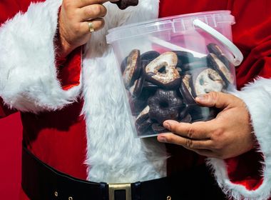  Ein Oberkörperfoto des Weihnachtsmannes, der einen klaren, runden Eimer mit dunklen und hellen Schoko-Lebkuchenbrezeln in der linken Hand hält. Mit der rechten Hand hebt er eine dunkel überzogene Brezel zum Mund. 