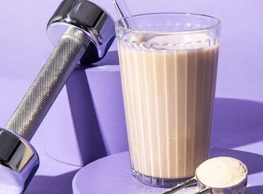 Ein Glas mit cremigem Vanille-Proteinshake und Glasstrohhalm steht auf einem lilafarbenen Podest, daneben ein Messlöffel mit veganem Vanille-Proteinpulver und eine silberne Hantel.