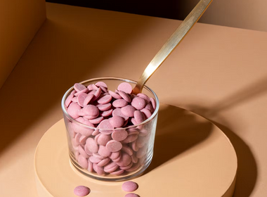 Rosafarbene Schoko-Drops aus Ruby-Kakaobohnen in einem kleinen Glas mit einem goldenen Löffel auf hellem Untergrund