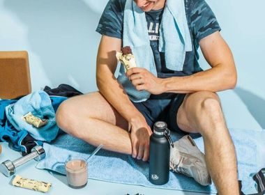 Ein Mann sitzt vor einem hellblauen Hintergrund auf einem Handtuch auf dem Boden. Er hat einen Proteinriegel in der Hand und vor ihm liegen diverse Proteinprodukte sowie Hanteln und eine Wasserflasche. Er selbst trägt Sportkleidung.