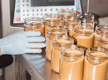 Auf einem Metalltisch in einem sterilen Lagerraum stehen Nussmusgläser – mit Inhalt, aber ohne Etikett. Eine Hand, die einen Einweghandschuh trägt, greift nach einem der Gläser.