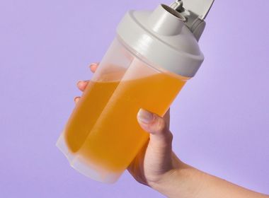 Hand hält einen mit Clear Protein halb gefüllten, durchsichtigen Proteinshaker mit grauem Deckel vor einem lilafarbenen Hintergrund.