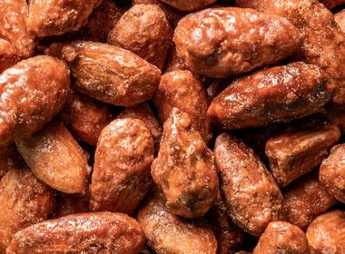 Ein detailliertes Nahaufnahmebild von einem Haufen gebrannter Mandeln. Sie sind alle mit einer glänzenden, karamellisierten Zuckerschicht überzogen und haben eine leicht unebene, knusprige Textur.