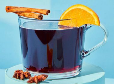 Ein Glas Glühwein oder Punsch, garniert mit einer Orangenscheibe und Zimtstangen, steht auf einem hellblauen Podest. Sternanis liegt daneben.