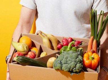  Ein Mann in einem weißen T-Shirt hält eine braune Pappkiste mit frischem Obst und Gemüse in der Hand. Die Kiste ist in mehrere Fächer unterteilt und enthält unter anderem Brokkoli, Bananen, Karotten, Paprika, Zucchini, Äpfel und Birnen. Der Mann steht vor einem leuchtend gelben Hintergrund.
