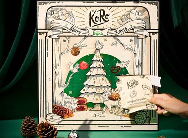 Vor einem grünen Hintergrund samt grünem Vorhang steht ein beigefarbener Adventskalender, auf dem ein gezeichneter Tannenbaum und Tiere zu sehen sind. Vor dem Kalender hält eine weibliche Hand ein Päckchen mit Laugenbrezeln in der Hand. Neben dem Kalender liegen Zapfen und ein Energy Ball.