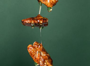 Drei goldbraune Pekannuskerne schweben in der Luft vor einem tiefgrünen Hintergrund. Sie sind mit klarem, glänzendem Karamell überzogen, das von der obersten Nuss über die mittlere bis zur untersten tropft. 