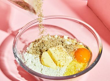 Eine Glas-Rührschüssel auf pinkem Hintergrund, gefüllt mit Backzutaten: Mehl, einem Ei, zwei Stücke Butter und eine helle Pulvermischung. Von oben wird weiteres bräunliches Pulver, vermutlich Proteinpulver, aus einem durchsichtigen Beutel in die Schüssel geschüttet.