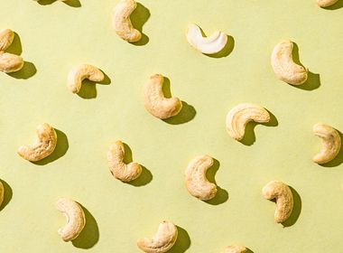 Einzelne Cashewkerne liegen in Reihen angeordnet auf einem hellgrünen Hintergrund. Die Cashewkerne sind ungeröstet und haben eine helle, cremeweiße Farbe. Sie werfen durch eine starke Lichtquelle von oben rechts Schatten.